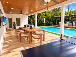 A poolside area with a wooden table and chairs, a ping pong table, and a view of a swimming pool surrounded by lush greenery. - The Quiet Luxury (Hollywood)