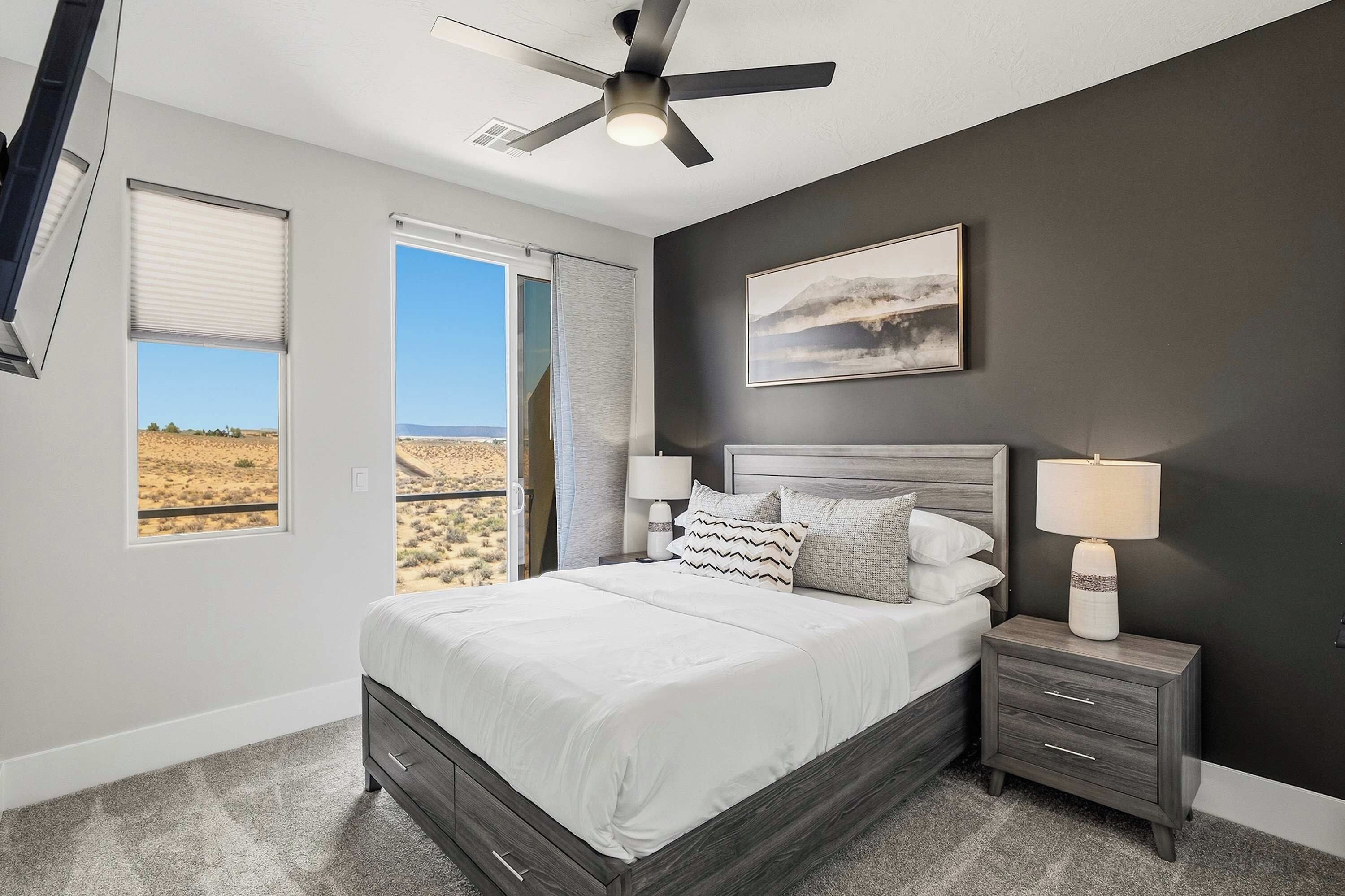 A bedroom with a bed, nightstands, a ceiling fan, a window with a view, and a painting on the wall.