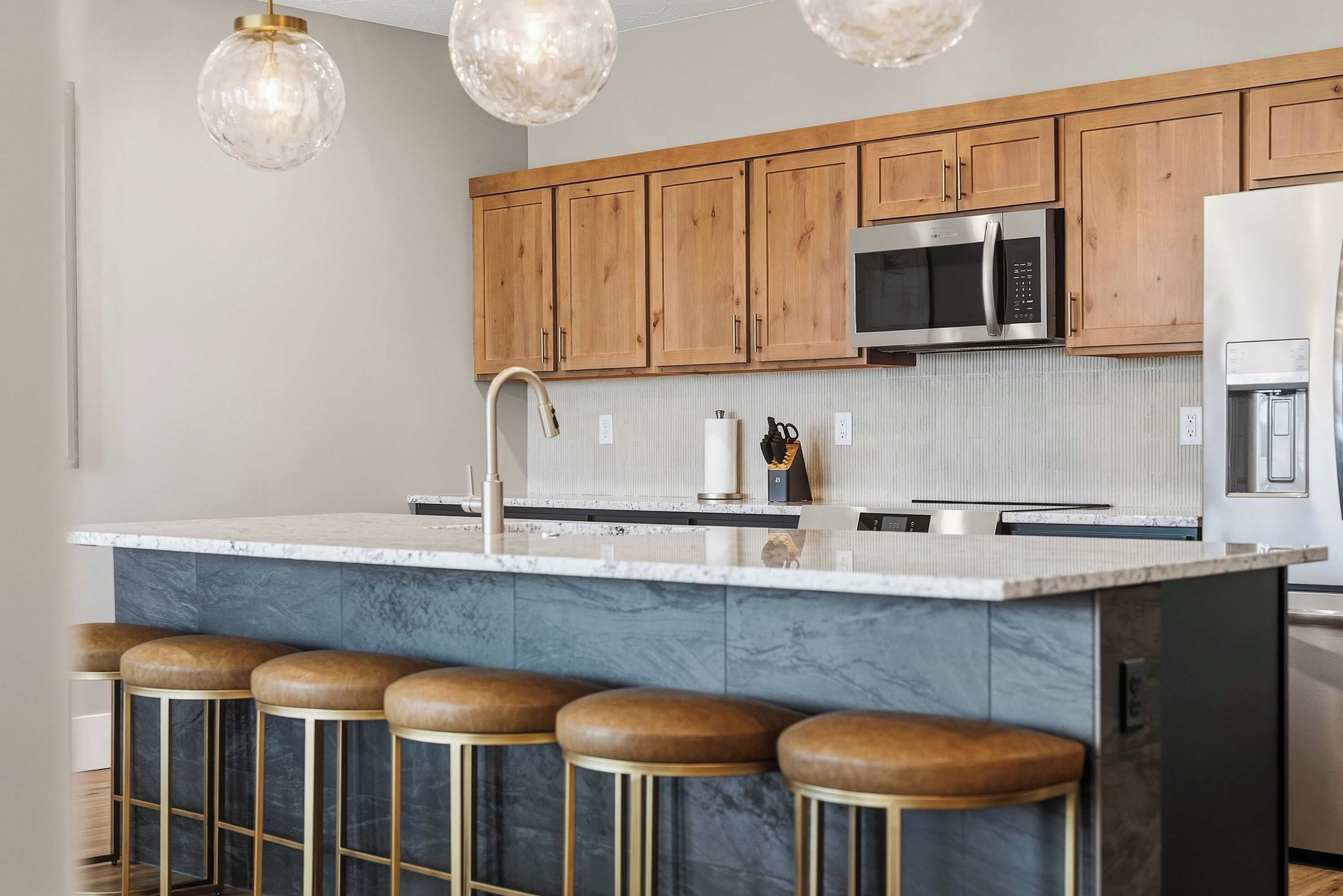 A modern kitchen with wooden cabinets, a stainless steel microwave, and a marble island with bar stools.