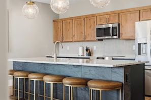 A modern kitchen with wooden cabinets, a stainless steel microwave, and a marble island with bar stools. - The Desert Terrace- Views, Sleeps 6, 3 BR (Canyon Point)