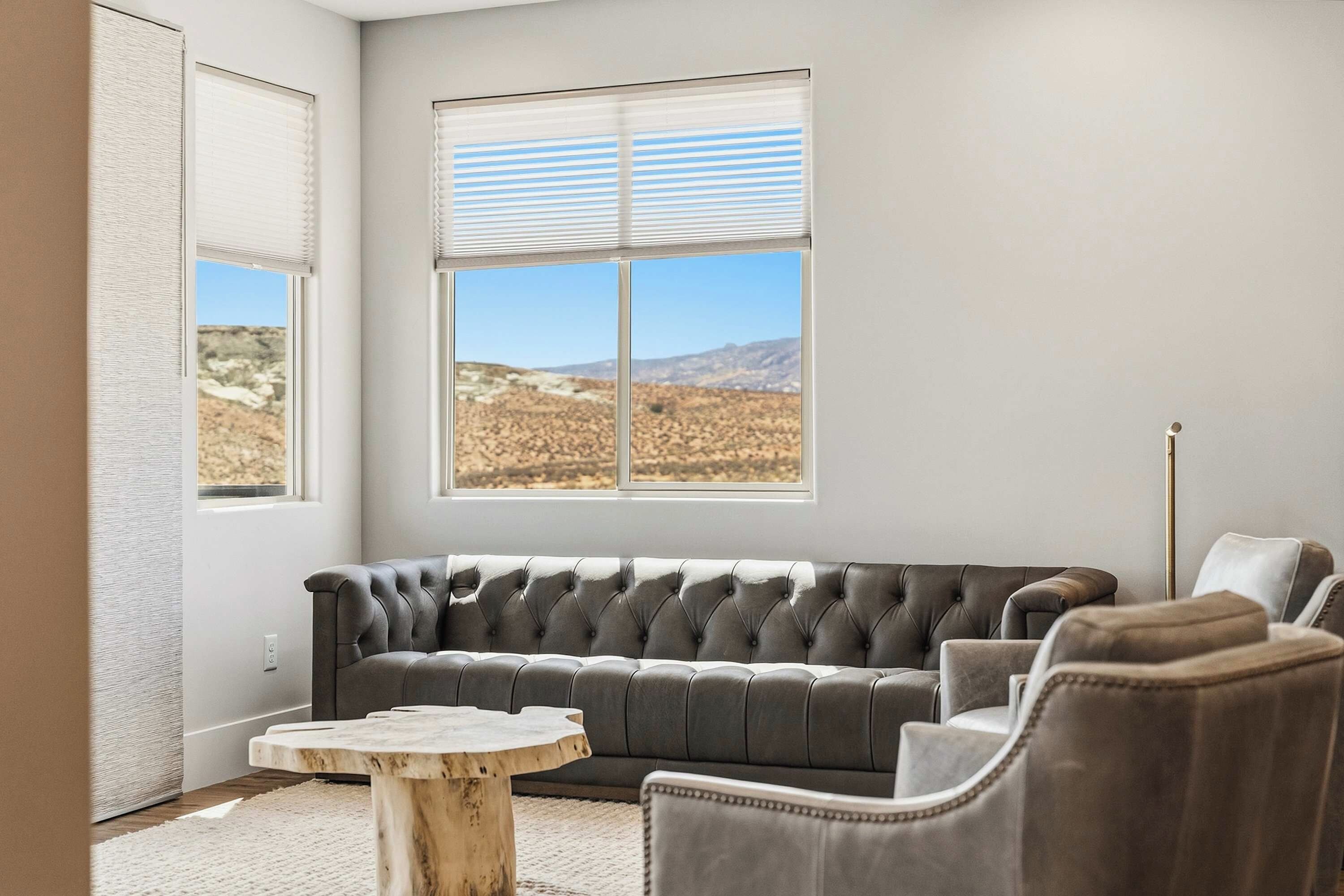 A modern living room with a grey sofa, a round wooden coffee table, and a view of a desert landscape through large windows.