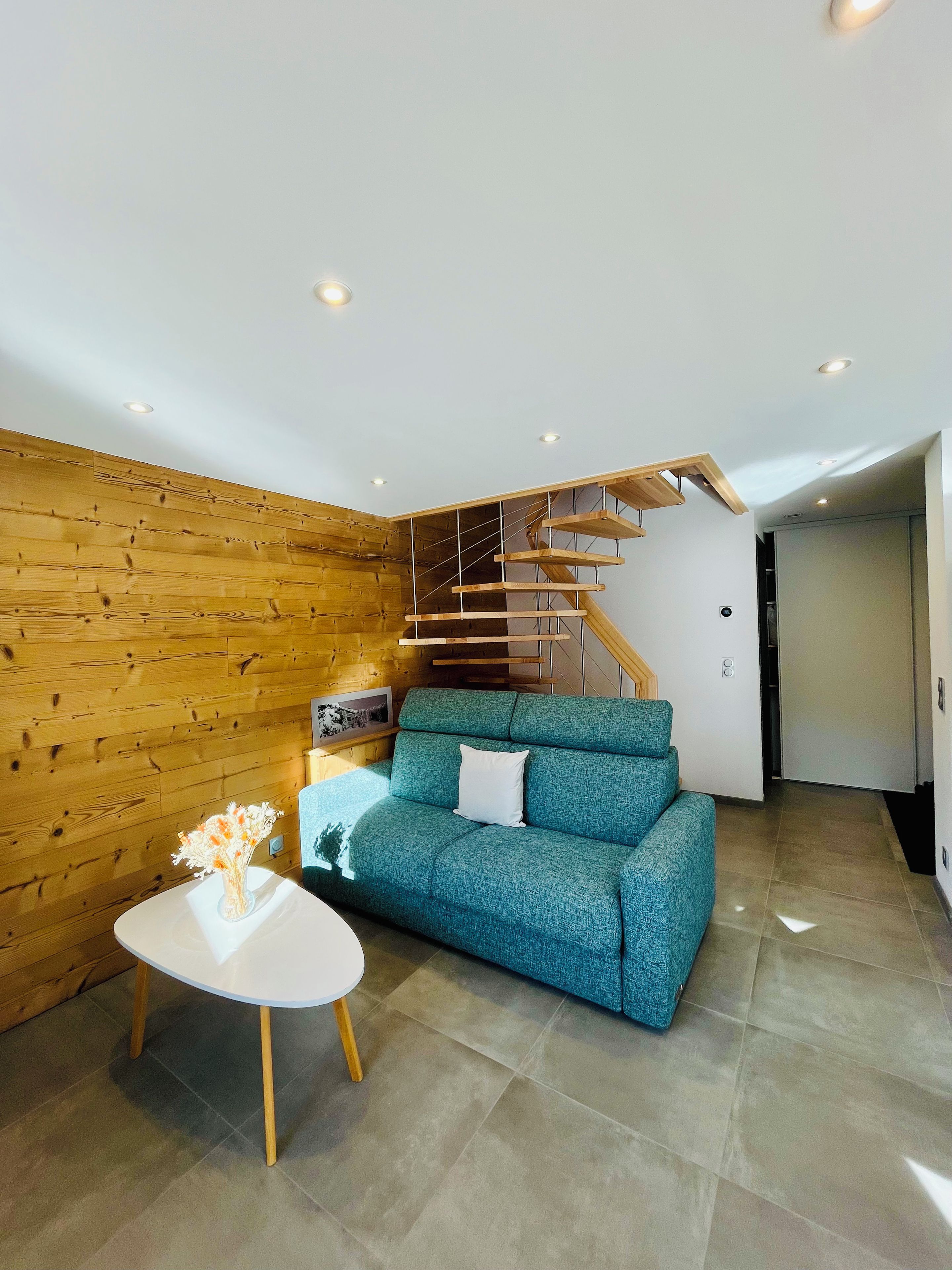 Una sala de estar moderna con una pared de madera, un sofá azul, una mesa blanca con un florero y una escalera de madera.