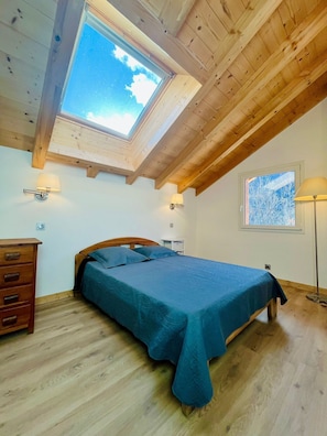 Ein Schlafzimmer mit einem Oberlicht, Holzbalken an der Decke, einem Bett mit blauer Bettdecke, einem Nachttisch, einer Kommode und einer Stehlampe.