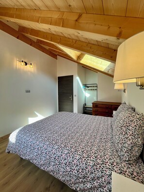 A bedroom with a sloped ceiling, a bed with a patterned bedspread, a wooden dresser, and a wall-mounted lamp. - New Listing (Brides-les-Bains)