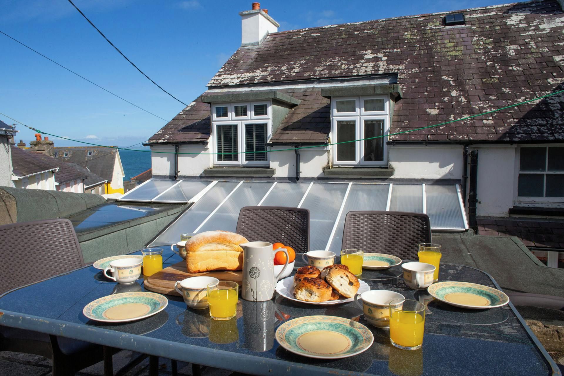 Une table à manger dressée avec des assiettes, des tasses et un pain, avec vue sur une maison et le ciel.