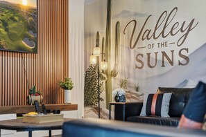 A modern living room with a large wall mural reading 'Valley of the Suns,' a wooden slat wall, a black leather sofa, a floor lamp, and a small table with a cactus and a plant. - Arizona Luxury Home. 5 Mins from Stadium. Play-Structure, Pool, Basketball Court (Peoria)