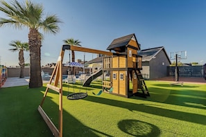 A wooden play structure with swings and a slide, surrounded by artificial grass and palm trees. - Arizona Luxury Home. 5 Mins from Stadium. Play-Structure, Pool, Basketball Court (Peoria)