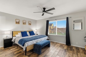 A bedroom with a bed, bedside tables, a ceiling fan, a window with curtains, and a view of buildings outside. - Arizona Luxury Home. 5 Mins from Stadium. Play-Structure, Pool, Basketball Court (Peoria)