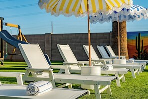 A row of white lounge chairs with umbrellas, a children's slide, and a mural on the wall. - Arizona Luxury Home. 5 Mins from Stadium. Play-Structure, Pool, Basketball Court (Peoria)
