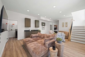A modern living room with a sectional sofa, a dining area with a kitchen in the background, and a staircase leading to an upper level. - Spacious Homes w/ Hot Tubs near Ark Encounter, Parking (Dry Ridge)