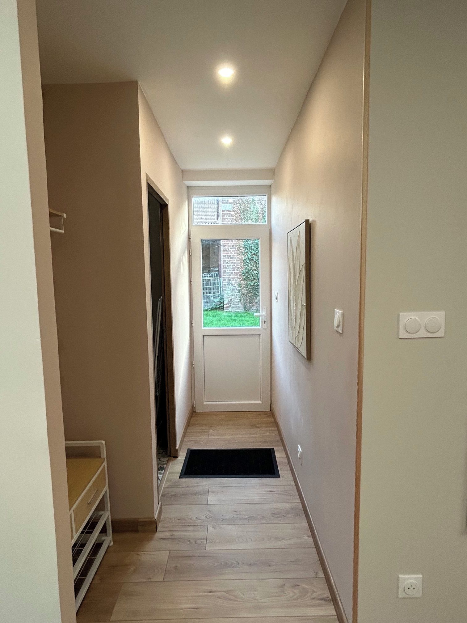 A narrow hallway with a door leading to a small room, a window with a view of greenery, and a black mat at the entrance.