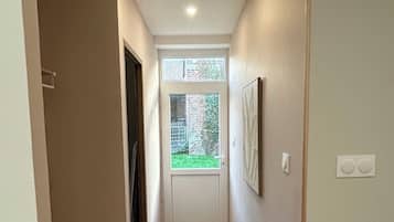 A narrow hallway with a door leading to a small room, a window with a view of greenery, and a black mat at the entrance.