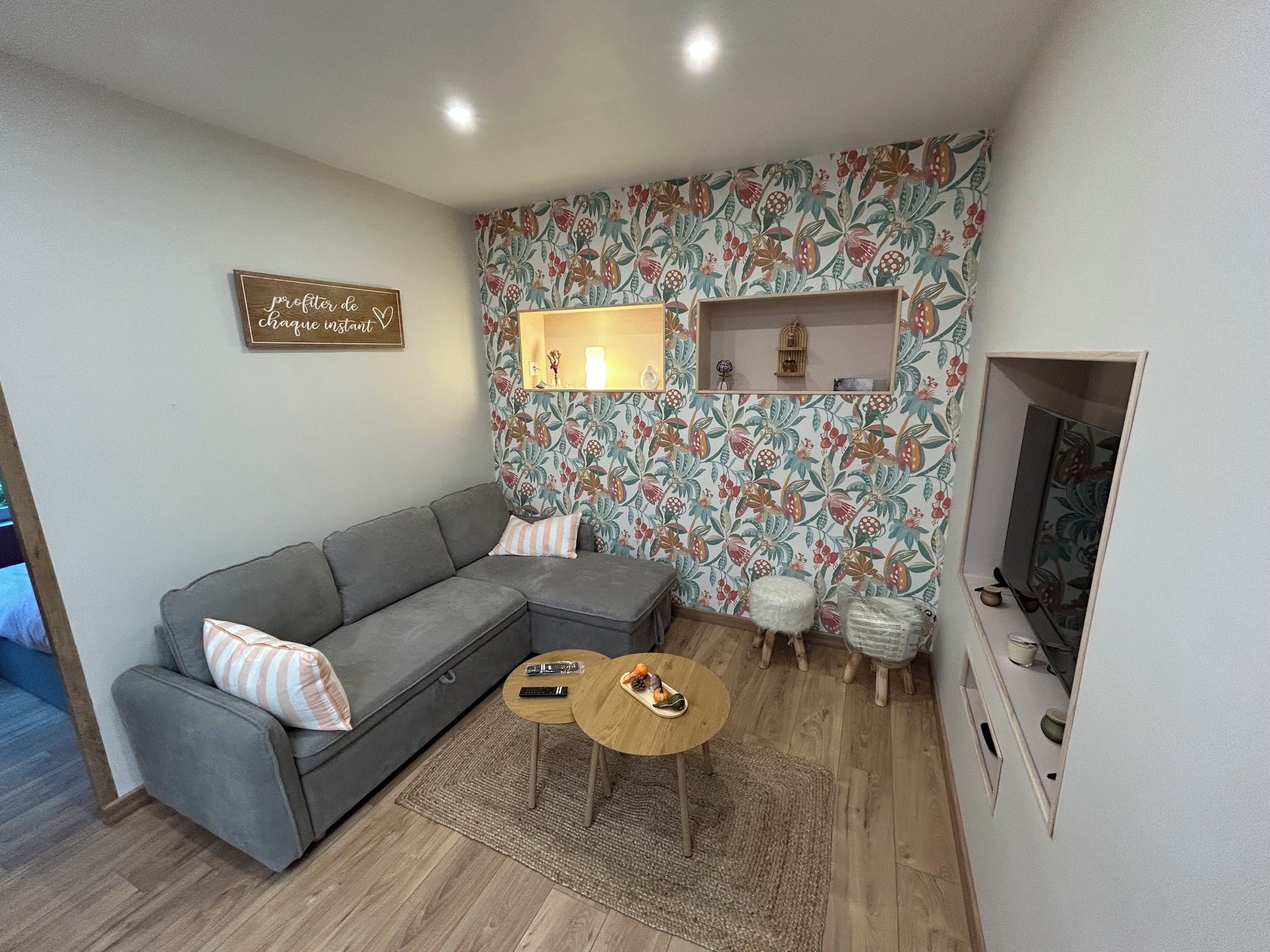 Una sala de estar con un sofá gris, dos mesitas redondas, un recamado decorativo con estampado floral y un televisor de pared.