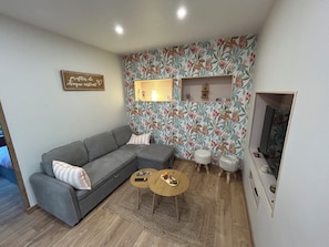 Una sala de estar con un sofá gris, dos mesitas redondas, un recamado decorativo con estampado floral y un televisor de pared.
