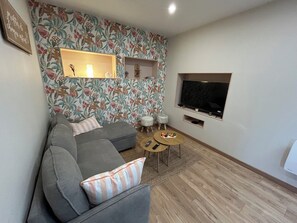 Una sala de estar con un sofá gris, una pequeña mesa redonda de centro, un televisor montado en la pared y un papel tapiz floral.