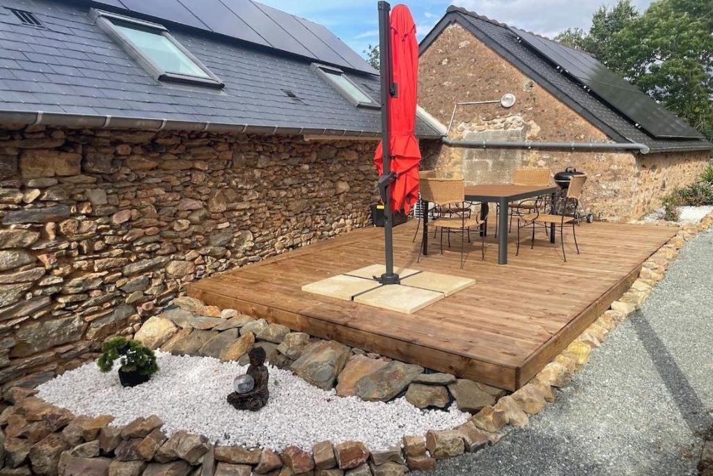 Un bâtiment en pierre avec une terrasse en bois, un parasol rouge et une table entourée de chaises.