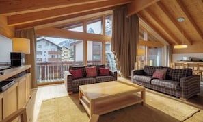 A cozy living room with two sofas, a coffee table, and a TV. Large windows offer a view of the outside. - MATTERHORN VIEW RETREAT – ELEGANT 4-BEDROOM ZERMATT APARTMENT (Zermatt)