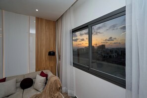A modern living room with a large window, a sofa with cushions, a floor lamp, and a view of a cityscape at sunset. - Premium one-bedroom apartment in Fann-Mermoz · Corniche within walking distance (Dakar)