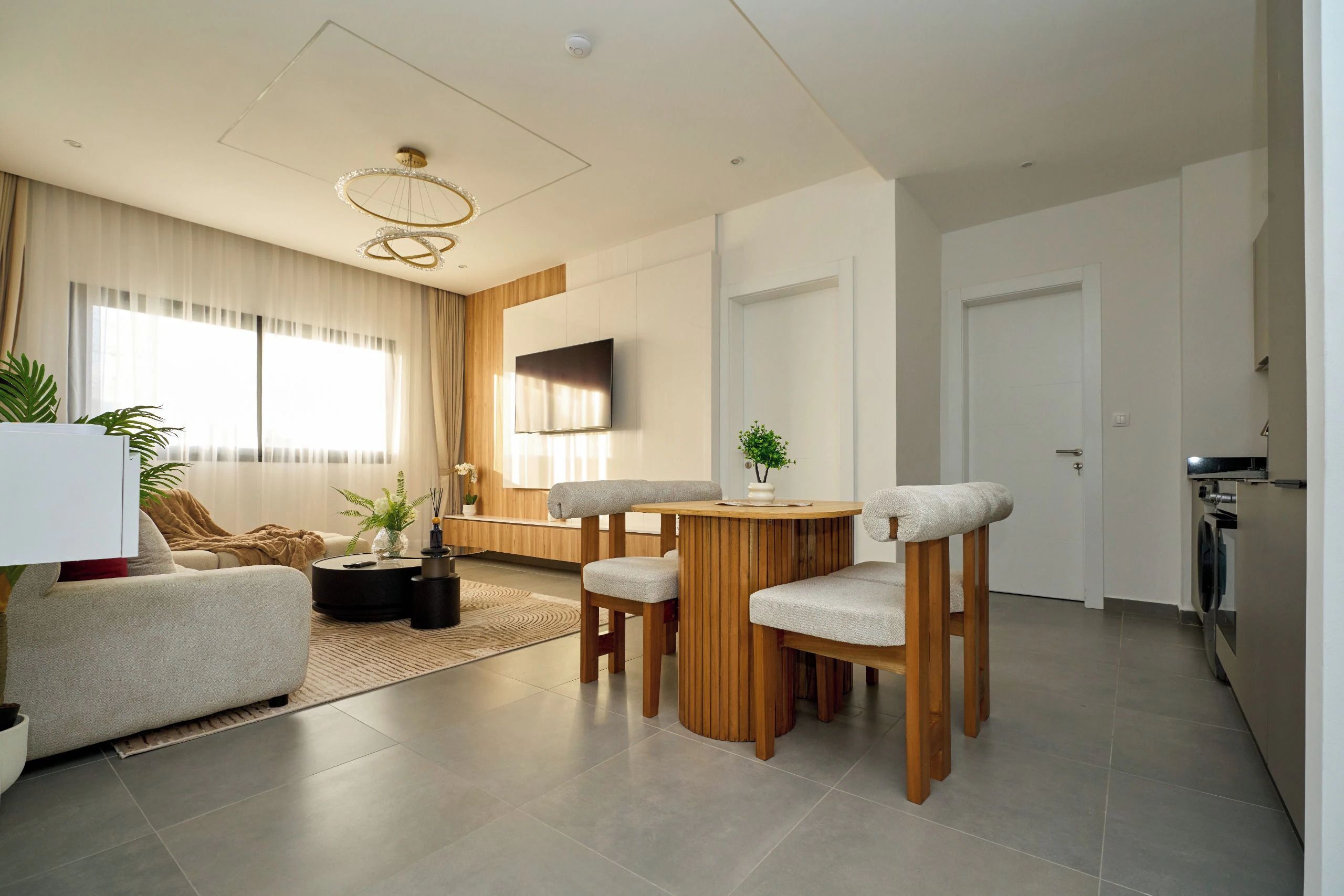 A modern living room with a sofa, coffee table, and chairs, a dining area with a table and chairs, a TV mounted on the wall, and a kitchen area with a washing machine.