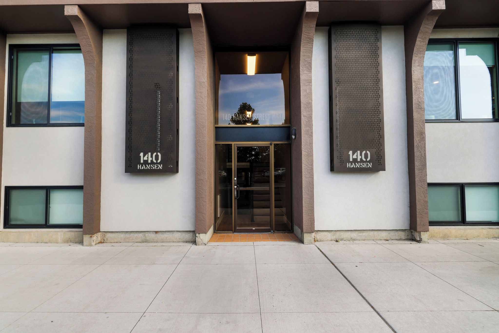 La façade d’un immeuble présente le numéro 140, affiché en caractères bien visibles sur deux panneaux verticaux, ainsi qu’une porte d’entrée en verre et un ciel dégagé que l’on peut apercevoir à travers les fenêtres.