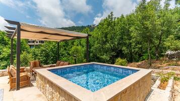 Piscina retangular com borda azulejada, rodeada por paredes de pedra, sob uma pérgola com toldo bege, com um pano de fundo de árvores verdes exuberantes.