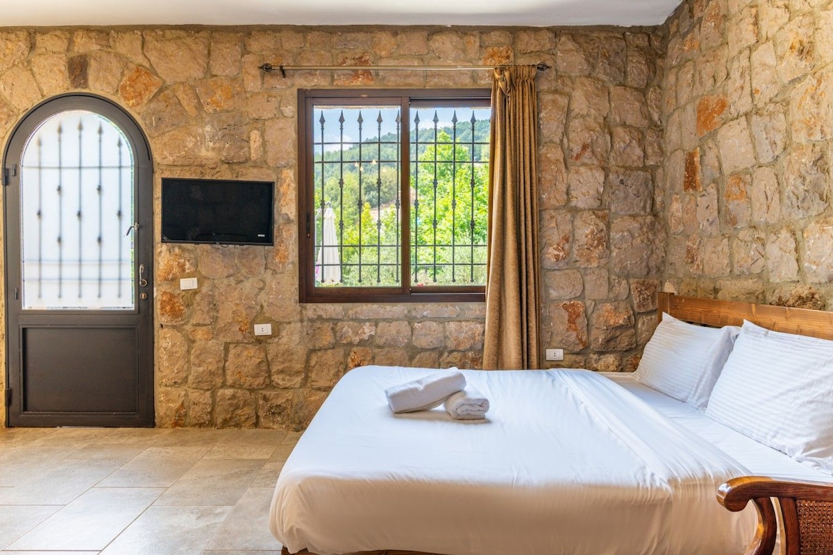 Quarto com parede de pedra, cama com roupa de cama branca, cabeceira de madeira, televisão fixada na parede e janela com vista para o verde.