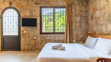 Habitación con pared de piedra, cama con ropa blanca, cabecero de madera, televisor montado en la pared y ventana con vista a follaje.