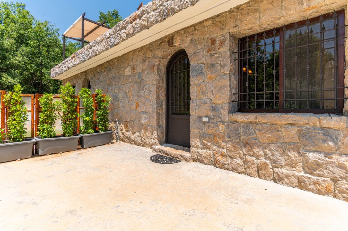 Um edifício de pedra com entrada em arco escura, uma janela com grades de metal e uma área de pátio coberta com vasos de plantas.