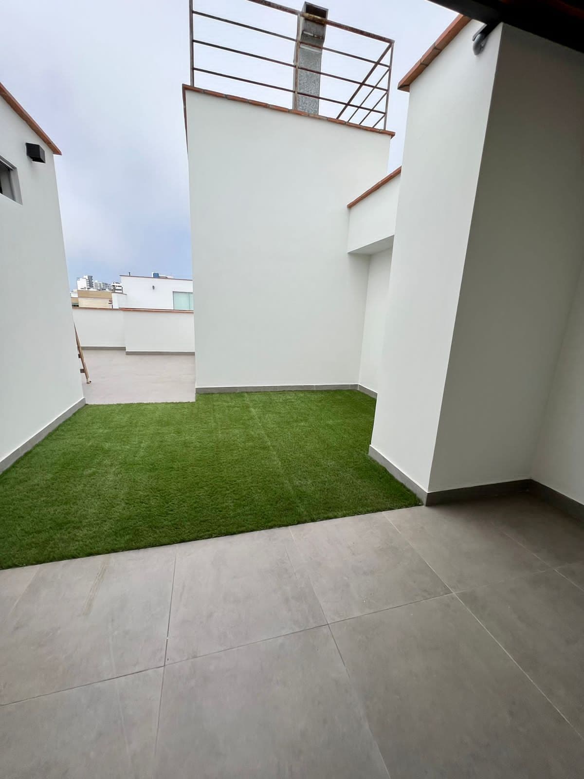 Una terraza en la azotea con un pequeño césped artificial, paredes blancas y barandilla de metal.