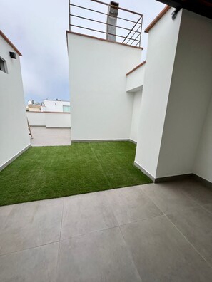 Una terraza en la azotea con un pequeño césped artificial, paredes blancas y barandilla de metal.