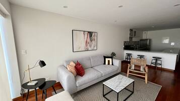 A modern living room with a grey sofa, a coffee table, and a dining area with bar stools.