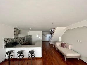 A modern kitchen with a central island, bar stools, and a staircase leading to a loft area with a sofa. - Walk to Larcomar | Peaceful 7th Floor Flat • AC (Miraflores)