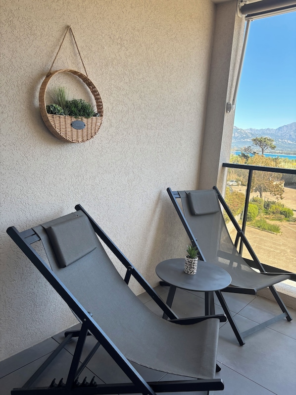 Un balcon avec deux fauteuils, une petite table et un panier suspendu en osier contenant des plantes vertes.