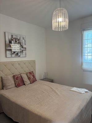 Une chambre à coucher avec une tête de lit capitonnée, un lit recouvert d’une couverture beige, des coussins à motifs, un luminaire suspendu et une œuvre d’art accrochée au mur.