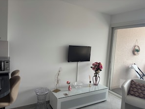 Ein modernes Wohnzimmer mit einem an der Wand befestigten Flachbildfernseher, einem Glastisch mit Dekorationsobjekten, einer Couch und einem Sessel.
