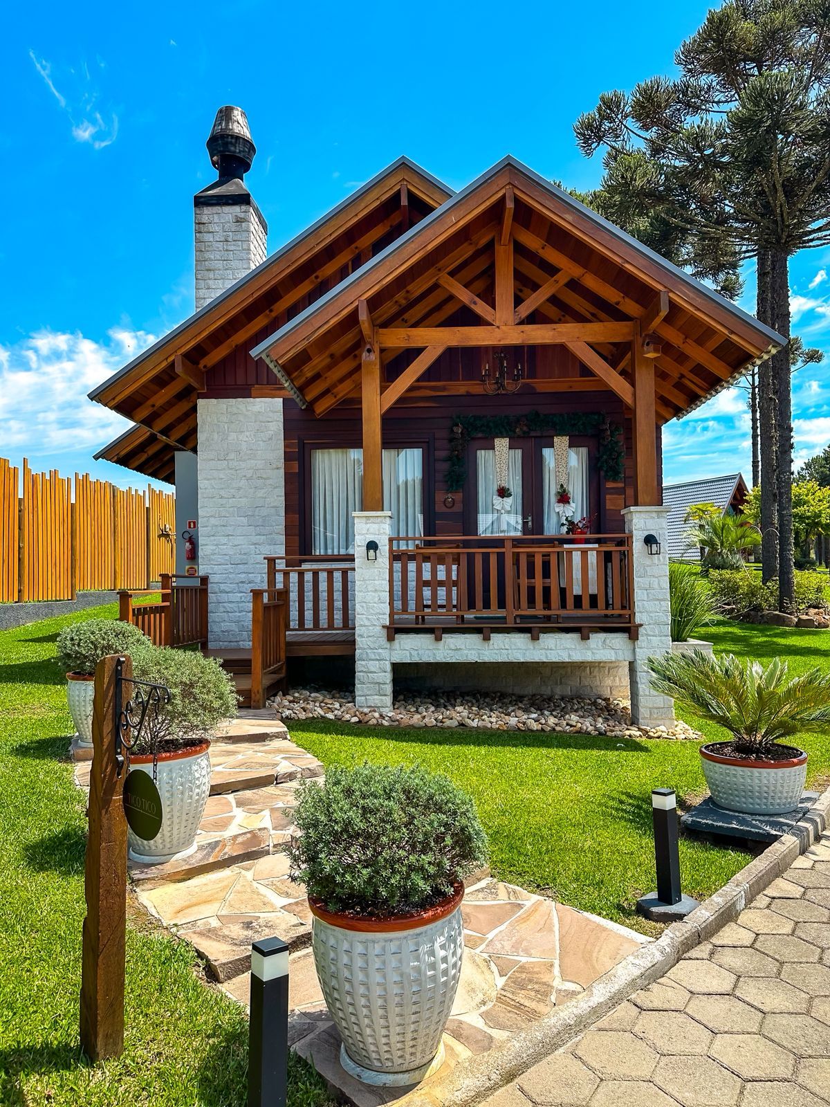 Une cabane en bois avec une cheminée en pierre, une véranda dotée de balustrades en bois et des plantes en pot au premier plan.