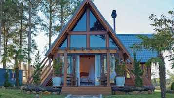 Una cabaña moderna de forma triangular con un amplio ventanal, terraza de madera y un camino de piedra que lleva a la entrada.