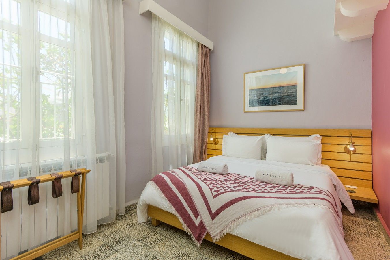 A hotel room with a bed, a window with sheer curtains, a wooden headboard, a wall-mounted picture, and a bedside lamp.