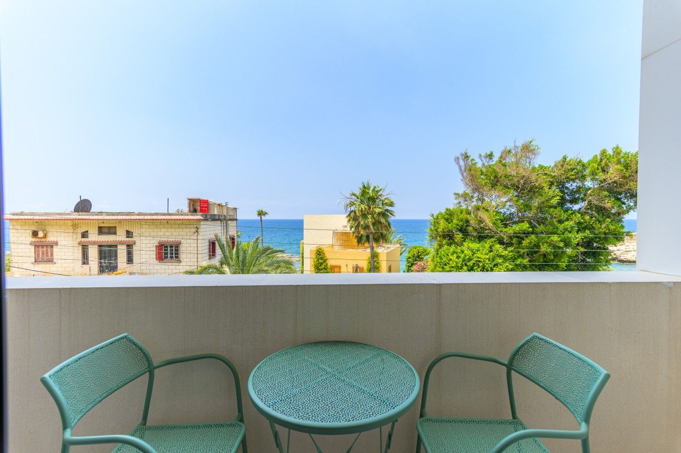 A balcony with two chairs and a small table, overlooking a coastal view with buildings and trees.