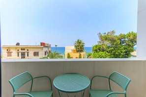 A balcony with two chairs and a small table, overlooking a coastal view with buildings and trees.