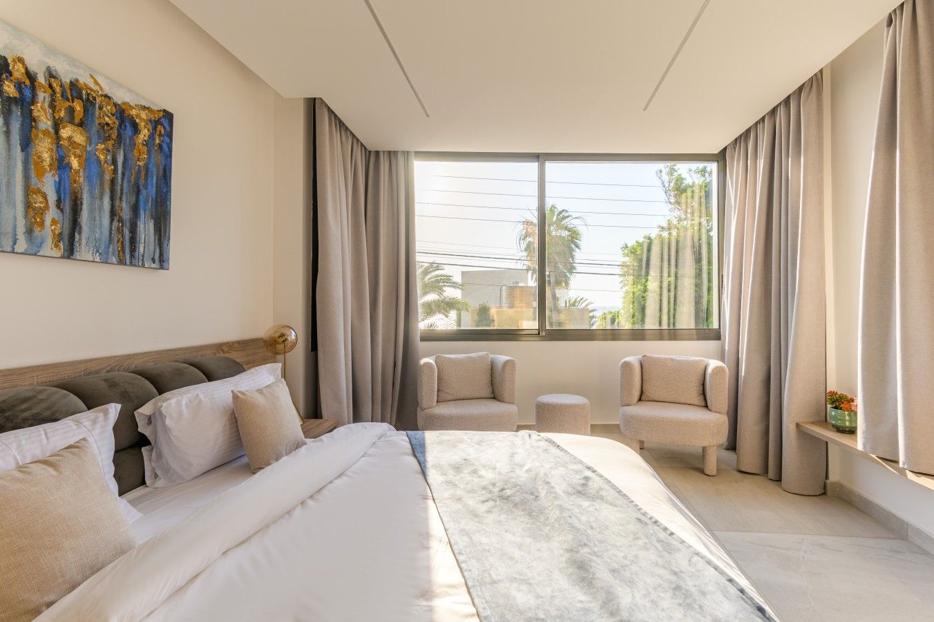 A modern bedroom with a large bed, two armchairs, a small table, and a large window with curtains.