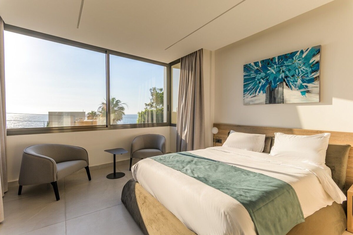Een hotelkamer met een bed, twee stoelen, een klein tafeltje, een groot raam met uitzicht op zee en een schilderij aan de muur.