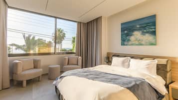 A hotel room with a large bed, two armchairs, a view of the sea, and a painting on the wall.