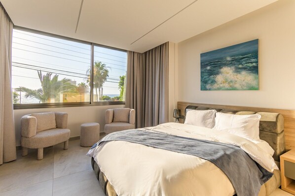 A hotel room with a large bed, two armchairs, a view of the sea, and a painting on the wall.