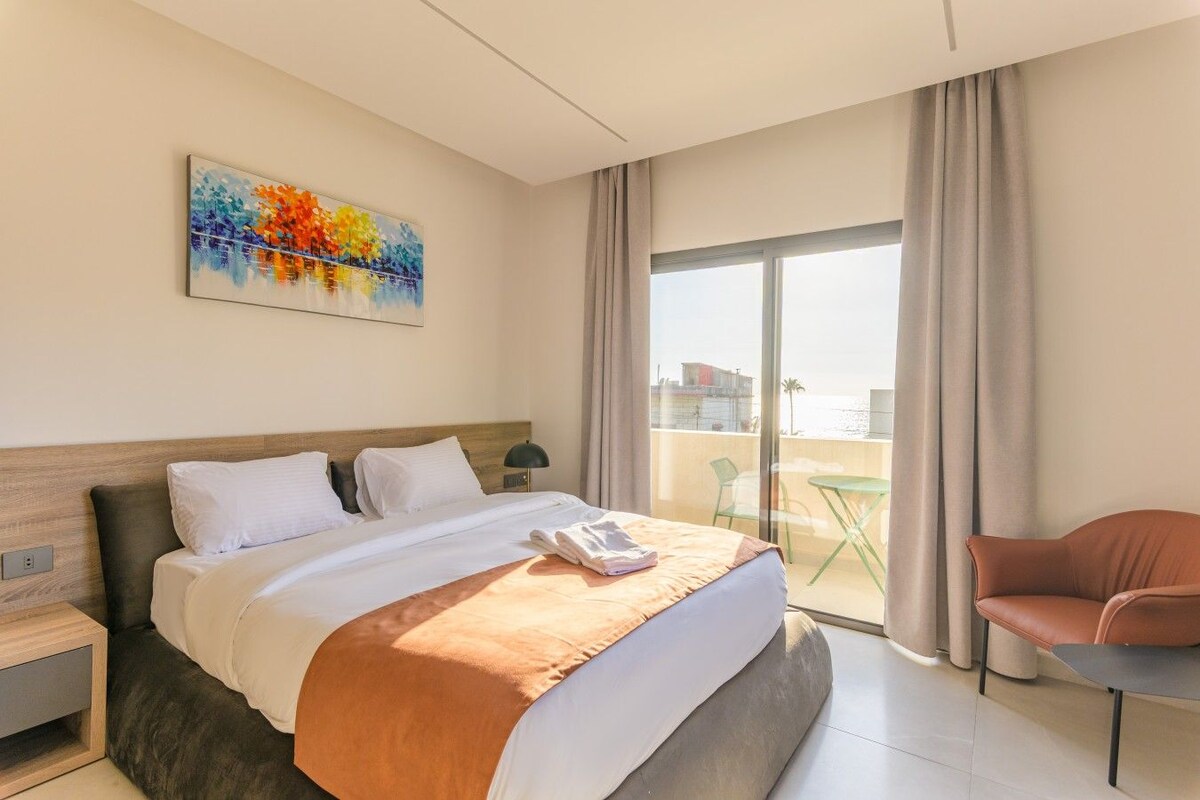 Een hotelkamer met een bed, een schilderij, een balkon met een stoel en uitzicht op zee.