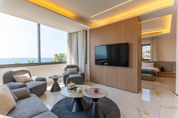 A modern hotel room with a large TV, comfortable seating, a view of the sea, and a balcony.
