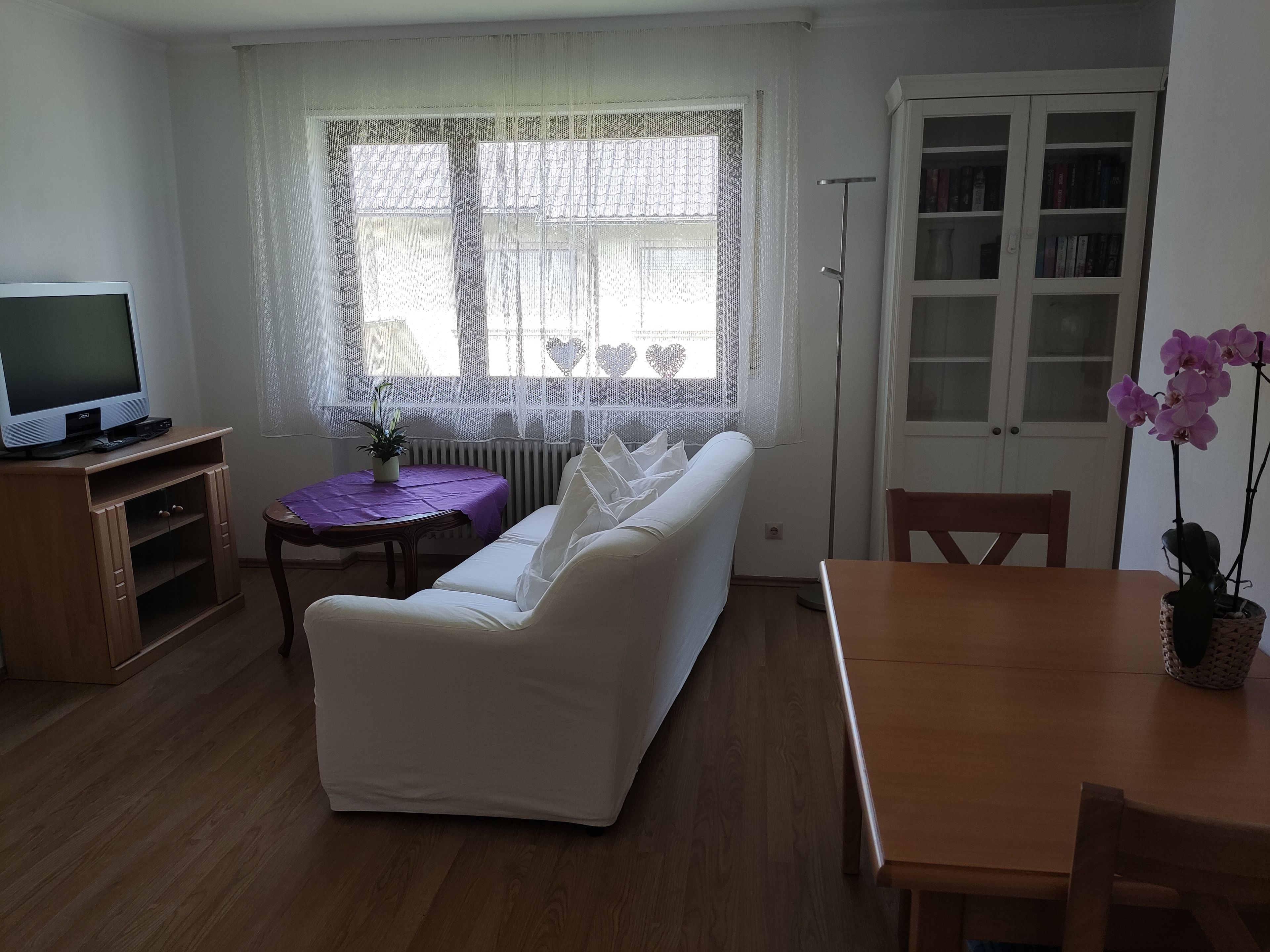 A living room with a white sofa, a small table with a purple tablecloth, a television on a wooden stand, and a bookshelf with books.