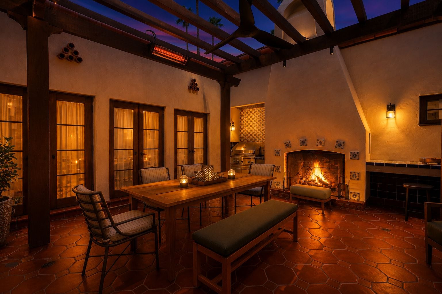 A cozy patio area with a wooden table, chairs, and a bench. There is a fireplace with a fire, and the space is illuminated by warm lighting.