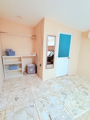 A room with a white tiled floor, a white desk with shelves, a grey storage bag, a blue basket, a mirror, and a door with a blue panel. - Cozy 1-bedroom apartment in delightful Machala with AC (Machala)