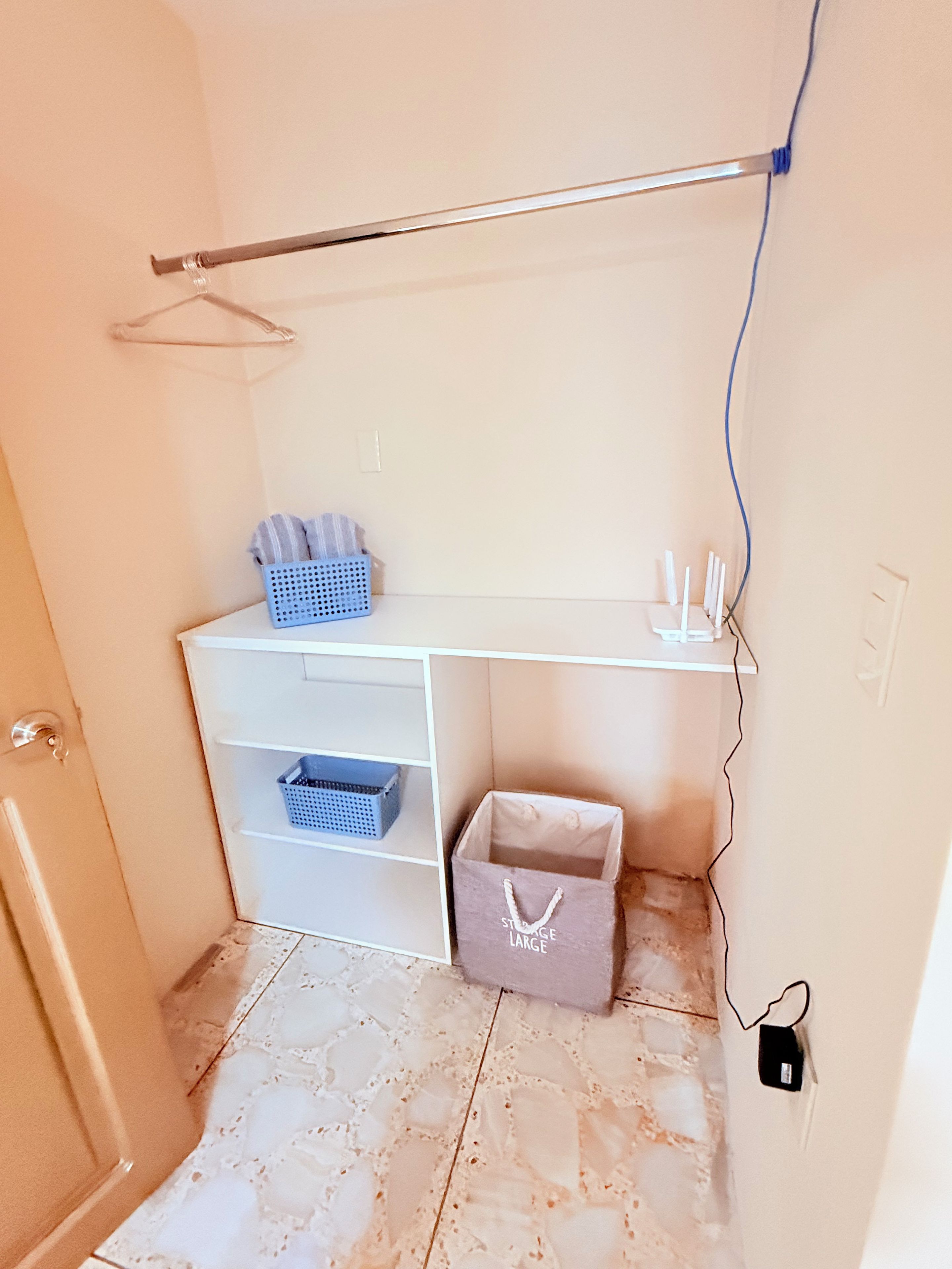 Une petite pièce, de taille modeste, avec un bureau blanc, une étagère suspendue, un panier bleu et une boîte de rangement grise.